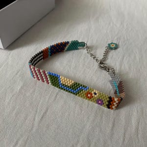 May include: A colorful beaded bracelet with a silver clasp and a small flower charm. The bracelet is made with a variety of colors, including green, blue, orange, and pink.