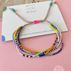 Puede incluir: Pulsera de cuentas multicolores con cierre de cordón tejido. La pulsera está hecha con pequeñas cuentas de colores y tiene un diseño simple y minimalista. La pulsera está atada con un cordón rosa y turquesa. La tarjeta en el fondo dice "Woo Den Boho" y "Handmade Miyuki designs by Burcu Aydil".