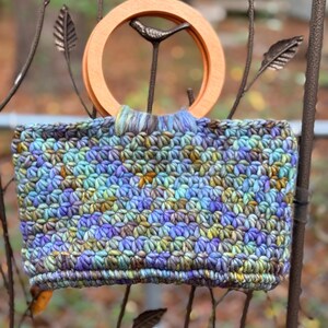 Könnte beinhalten: Eine handgefertigte Häkeltasche mit Holzgriff. Die Tasche hat ein strukturiertes Stichmuster in Blau-, Lila-, Grün- und Gelbtönen. Der Griff hat eine helle, braune, kreisförmige Form. Die Tasche hängt an einer dekorativen Metallstruktur.