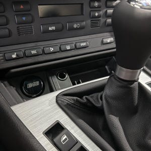 May include: Interior view of a car's center console. The image shows a black gear shift, a brushed metal panel, and various buttons and controls. A circular button with the text "E46" is visible.