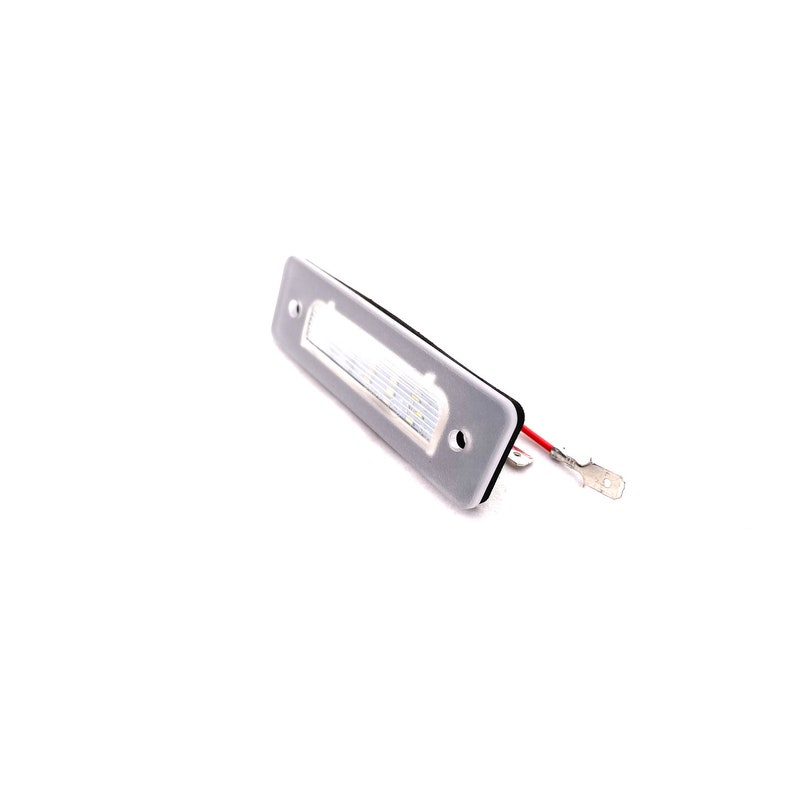 May include: A white LED license plate light with two wires attached. The light is rectangular and has a gray plastic housing.