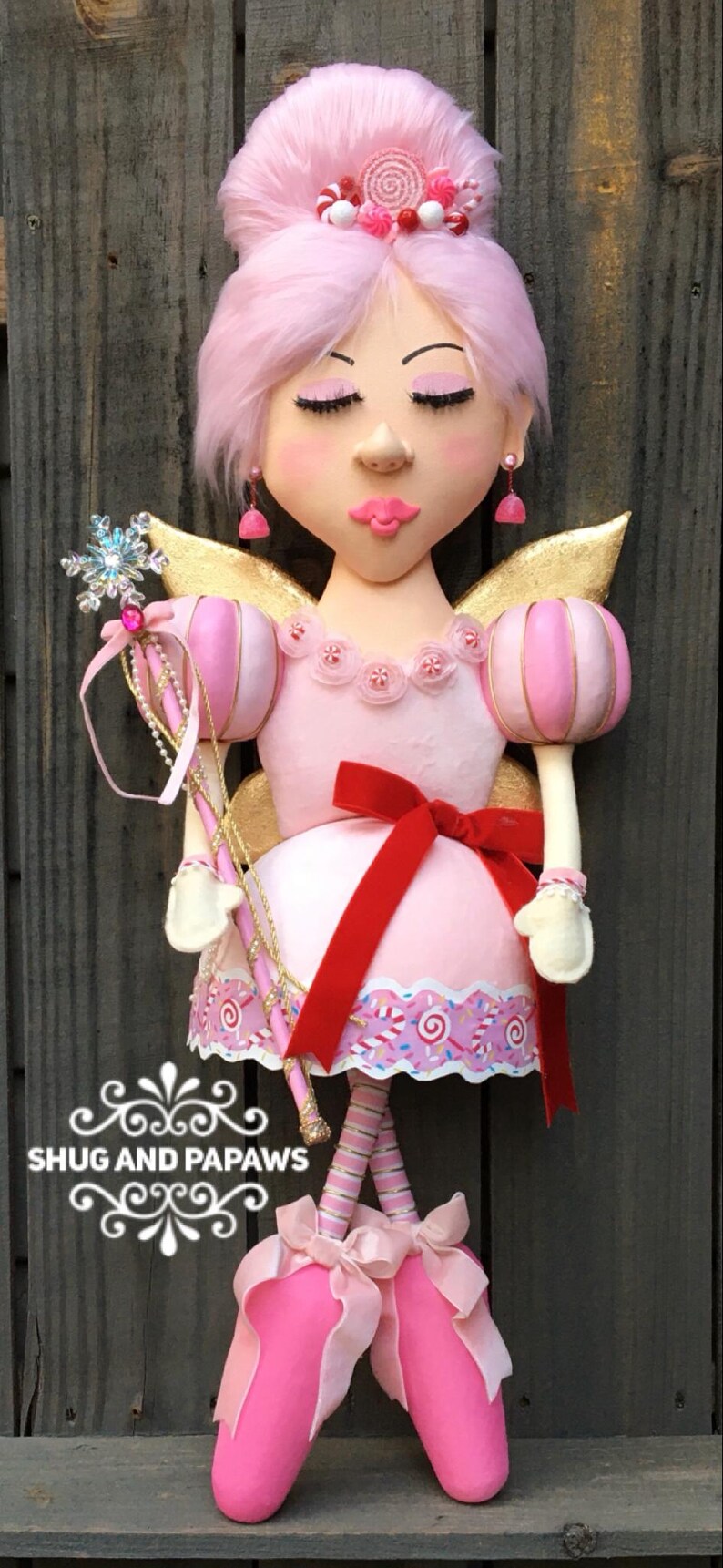 May include: A whimsical fairy doll with pink hair, a pink dress, and gold wings. The doll holds a snowflake wand and has a red bow. The text "SHUG AND PAPAWS" is visible.