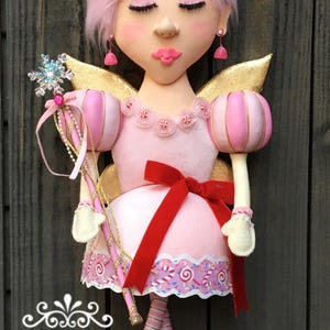 May include: A whimsical pink fairy doll with closed eyes, gold wings, and a red bow. The doll has a pink dress with a candy-themed trim, a snowflake wand, and pink ballet slippers. The doll's hair is styled with a pink bun and candy accents.