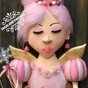 May include: A whimsical fairy figurine with pink hair, rosy cheeks, and closed eyes. The fairy wears a pink dress with candy accents, gold wings, and a red bow. A snowflake wand and the text "Shug and Papaws" are also visible.