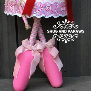 May include: A whimsical pink ballerina doll with a red sash and gold accents. The doll's legs are striped pink and silver, and it wears pink ballet slippers with large pink bows. The text "SHUG AND PAPAWS" is visible.