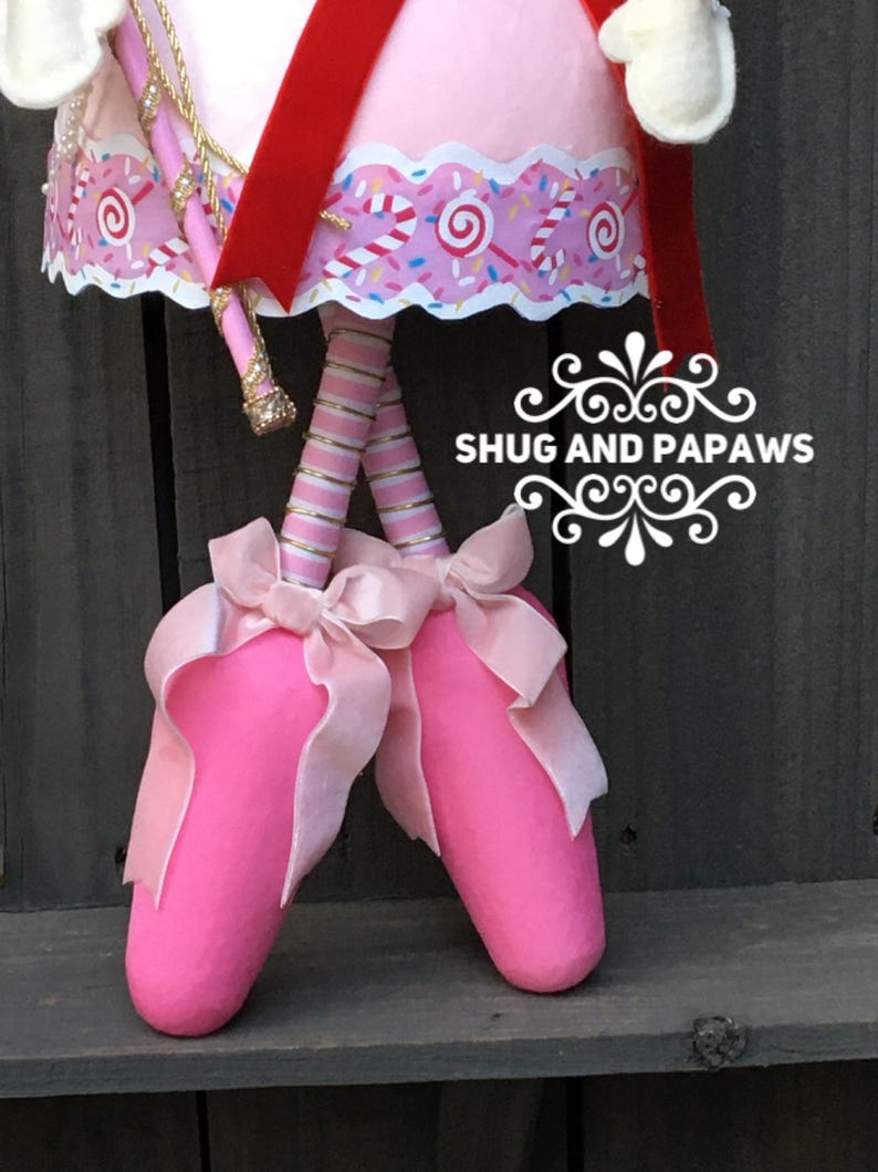 May include: A whimsical pink ballerina doll with a red sash and gold accents. The doll's legs are striped pink and silver, and it wears pink ballet slippers with large pink bows. The text "SHUG AND PAPAWS" is visible.
