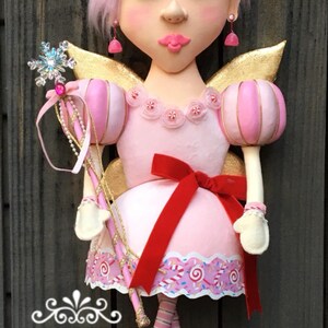 May include: A whimsical fairy doll with pink hair, a pink dress, and gold wings. The doll holds a snowflake wand and has a red bow. The text "SHUG AND PAPAWS" is visible.