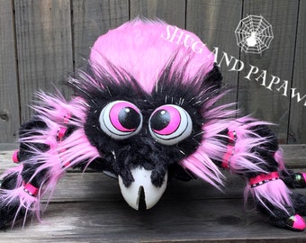 May include: A plush spider toy featuring long, shaggy black and bright pink faux fur, large round eyes with pink pupils, and white fangs. The text SHUG AND PAPAWS with a spiderweb icon is visible in the upper right corner of the image.