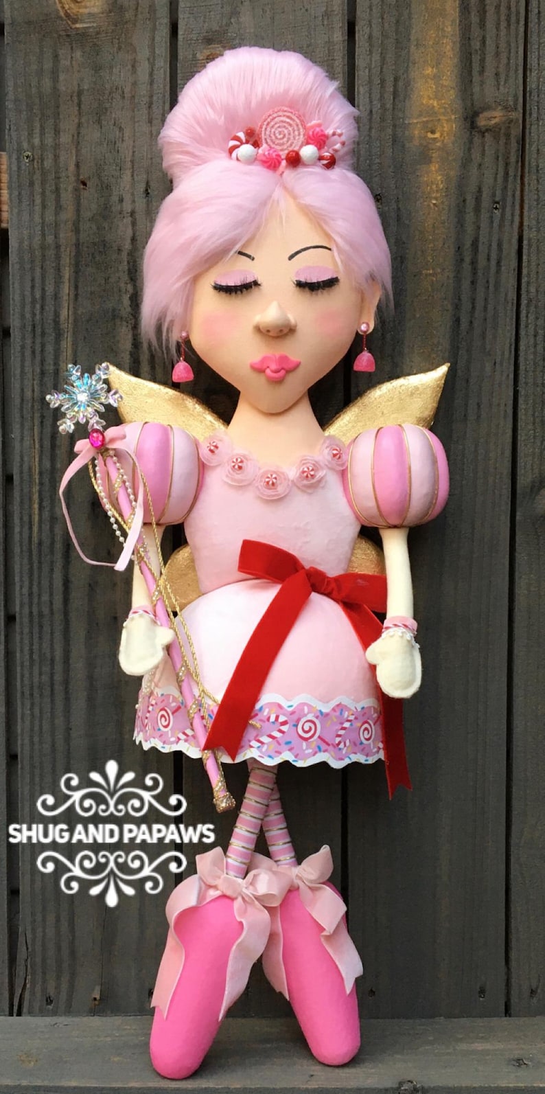 May include: A whimsical pink fairy doll with closed eyes, gold wings, and a red bow. The doll has a pink dress with a candy-themed trim, a snowflake wand, and pink ballet slippers. The doll's hair is styled with a pink bun and candy accents.