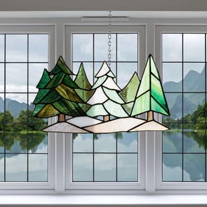 Könnte beinhalten: Glasfenster-Kunstwerk mit einer Waldszene mit immergrünen Bäumen in Grün- und Weißtönen. Das Kunstwerk wird von einer Kette gehalten und hängt vor einem Fenster mit Blick auf einen See und Berge.