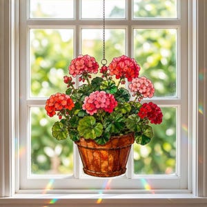 Puede incluir: Un atrapasoles de vidrieras que representa una planta de geranio en maceta con racimos de flores rojas y rosas vibrantes y hojas verdes, suspendido por una cadena de metal. La pieza presenta una maceta marrón texturizada ante una ventana.