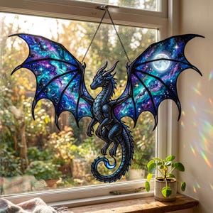 以下が含まれることがあります： ステンドグラスのドラゴンの窓飾り。ドラゴンは主に青色で、複雑なディテールが施されています。その翼は、青、紫、白の色合いの銀河のデザインを特徴としています。満月が片方の翼に見えます。ドラゴンはチェーンで吊り下げられています。