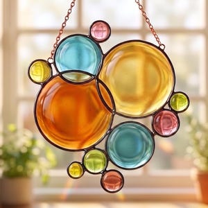 Op de afbeelding: Een gebrandschilderd raamdecoratie met een bubbelontwerp. Het glas is in tinten oranje, geel, blauw, groen en roze, met een donkerbruin metalen frame en een koperen ketting om op te hangen. Ronde decoratie.