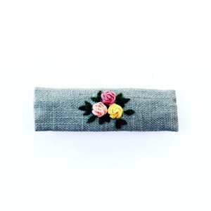 May include: A grey fabric rectangle with a small embroidered floral design featuring three roses in pink, yellow, and peach colours.