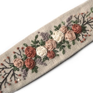Op de afbeelding: Een beige linnen lint met geborduurde bloemen in tinten roze, wit en bruin. De bloemen zijn in een rij langs het lint gerangschikt, met bladeren en stelen ertussen.