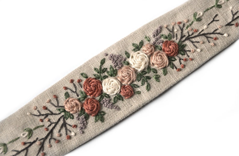 Op de afbeelding: Een beige linnen lint met geborduurde bloemen in tinten roze, wit en bruin. De bloemen zijn in een rij langs het lint gerangschikt, met bladeren en stelen ertussen.