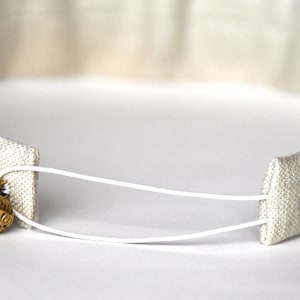 Op de afbeelding: Een wit hoofdband met een wit elastiek en een gouden metalen knoop. Het hoofdband is gemaakt van een zachte, geweven stof.