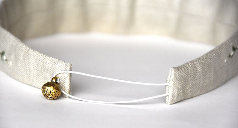 Op de afbeelding: Een wit hoofdband met een wit elastiek en een gouden metalen knoop. Het hoofdband is gemaakt van een zachte, geweven stof.