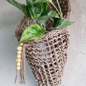 May include: A hanging planter made of natural woven material with a light brown colour. The planter is filled with a green and white variegated plant. The planter has a decorative wooden bead garland hanging from it.