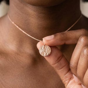 May include: A delicate gold-colored necklace featuring a textured, circular pendant. The pendant hangs from a fine chain, creating a minimalist and elegant design. The necklace is being held up by a hand.