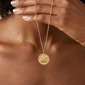 May include: A gold-colored necklace with a circular pendant. The pendant has a textured surface with a design. The necklace is being held by a person.