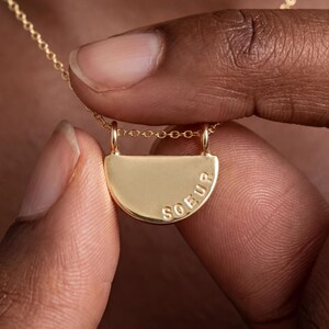 A person's fingers holding a gold-colored necklace with a pendant engraved with the word "SOEUR".
