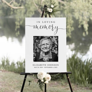May include: A white sign with black text that reads "In loving memory" with a black and white photo of an older woman. The text below the photo reads "Elizabeth Johnson, May 20, 1930 - September 4, 2025".