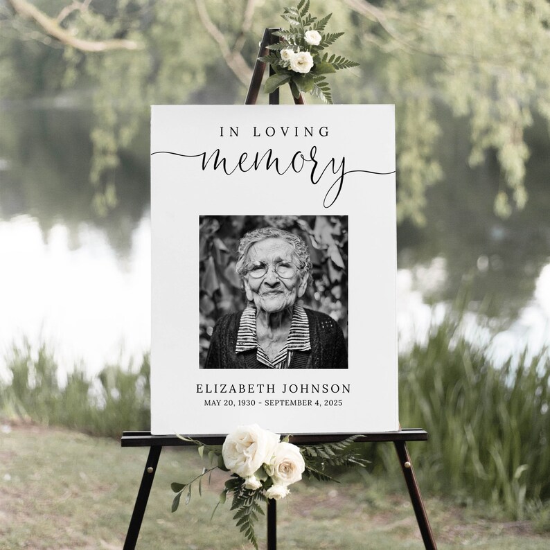 May include: A white sign with black text that reads "In loving memory" with a black and white photo of an older woman. The text below the photo reads "Elizabeth Johnson, May 20, 1930 - September 4, 2025".