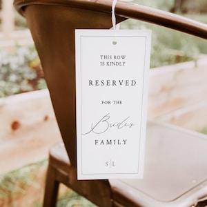 May include: White sign with black text that reads "This row is kindly reserved for the Bride's Family". The sign is hanging from a chair.