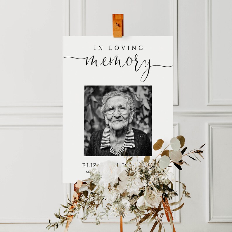 May include: A black and white photo of an older woman with glasses is on a white sign with the text "In loving memory". The sign is on a wooden easel with white flowers.