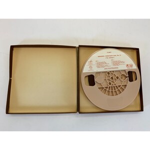 May include: A brown box with a beige interior, containing a reel-to-reel audio tape. The tape is labelled "Prokofiev:  'Classical' Suite, Op. 31  "The Buffoon'" and has a red and white label.