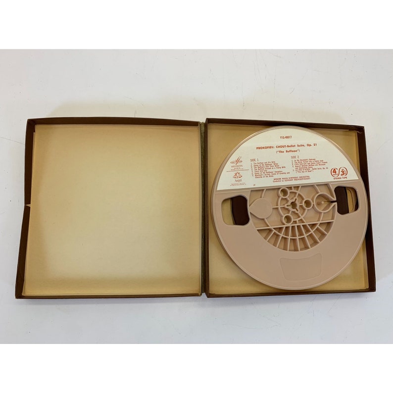 May include: A brown box with a beige interior, containing a reel-to-reel audio tape. The tape is labelled "Prokofiev:  'Classical' Suite, Op. 31  "The Buffoon'" and has a red and white label.