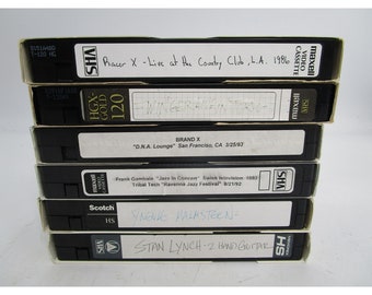 May include: A stack of VHS tapes with handwritten labels. The labels include the names of bands and artists such as Racer X, Winger, Brand X, Frank Gambale, Yngwie Malmsteen, and Stan Lynch. The tapes are labeled with the date and location of the recordings.