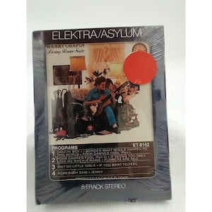 May include: A blue 8-track cassette tape case with a photo of a man playing a guitar in a living room. The case has the title "Living Room Suite" and the artist "Garry Chapin". The case also has the title "Elektra/Asylum" and the track list: "Dancin Boy", "Poor Damned Fool", "Why Do Little Girls", and "Somebody Said".