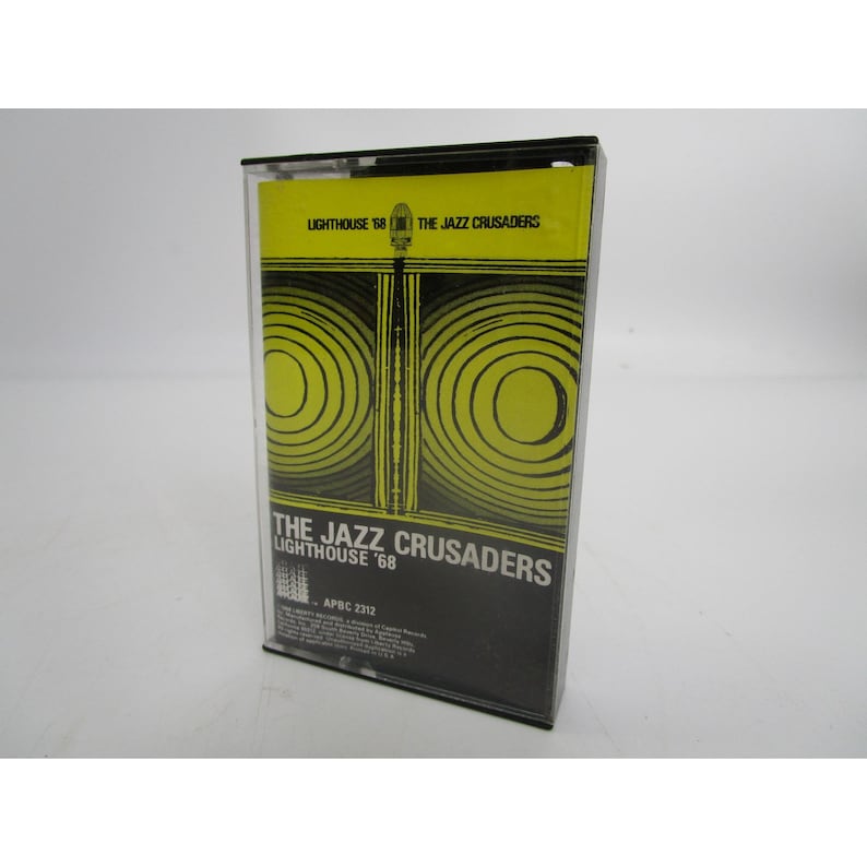 Op de afbeelding: Een zwarte cassetteband met een geel en zwart ontwerp. De band is gelabeld met "The Jazz Crusaders Lighthouse '68" en "APBC 2312".