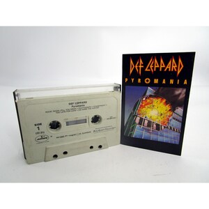 May include: A white cassette tape with a black label. The label has the text "DEF LEPPARD Pyromania" and a list of songs. The cassette tape is next to a black and white album cover with the text "DEF LEPPARD PYROMANIA".