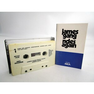 May include: A white cassette tape with the title "James Gang Rides Again" and the artist "James Gang" printed on the label. The cassette tape is in a plastic case with a clear window to see the tape. The case also has the MCA Records logo printed on it. The cassette tape is next to a white paper booklet with the title "James Gang Rides Again" printed in black.