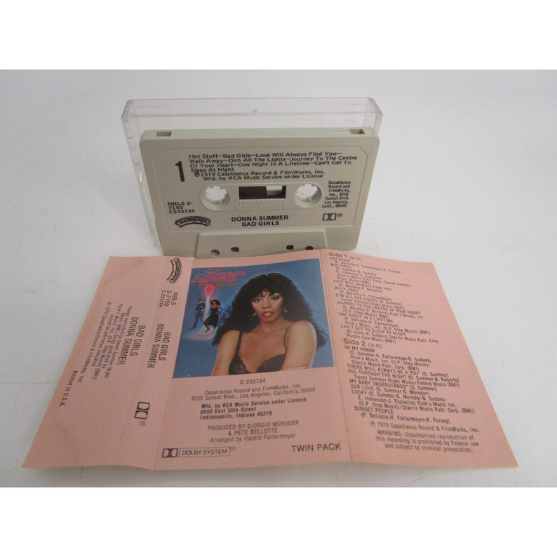 May include: A cassette tape case with a white background and a picture of Donna Summer on the front. The case is labeled "Donna Summer Bad Girls" and includes the text "Hot Stuff - Bad Girls - Love Will Always Find You - Dim All The Lights - Journey To The Centre Of Your Heart - One Night In A Lifetime - Can't Get To You - Walk Away - Dim All The Lights".