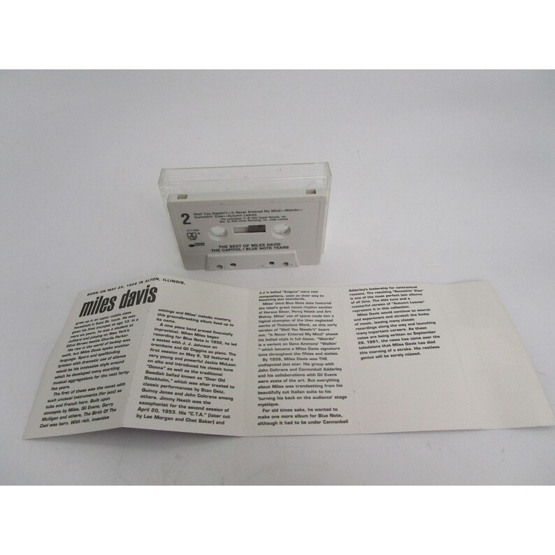 Op de afbeelding: Een witte cassettebanddoos met een witte cassetteband erin. De cassetteband is gelabeld met "The Best of Miles Davis - The Capitol / Blue Note Years". De doos bevat ook een wit boekje met tekst over Miles Davis.
