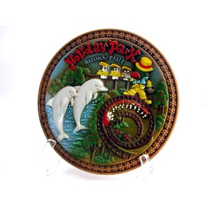 Op de afbeelding: Een bruin keramiek bord met een reliëfdesign van een achtbaan, twee dolfijnen en een clown. Het bord heeft de tekst "Holiday Park Hassloch/Pfalz" in een bruin, geel en rood lettertype.