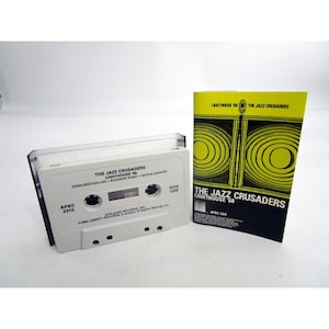 Op de afbeelding: Een witte cassetteband met de titel "The Jazz Crusaders Lighthouse '68" en de songtitels "Dooga-Boo-Ga-Loo", "Eleanor Rigby" en "Native Dancer". De cassetteband zit in een doorzichtige plastic hoes met een zwart label. De cassetteband ligt naast een zwart en geel papieren inlegvel met dezelfde titel en bandnaam.