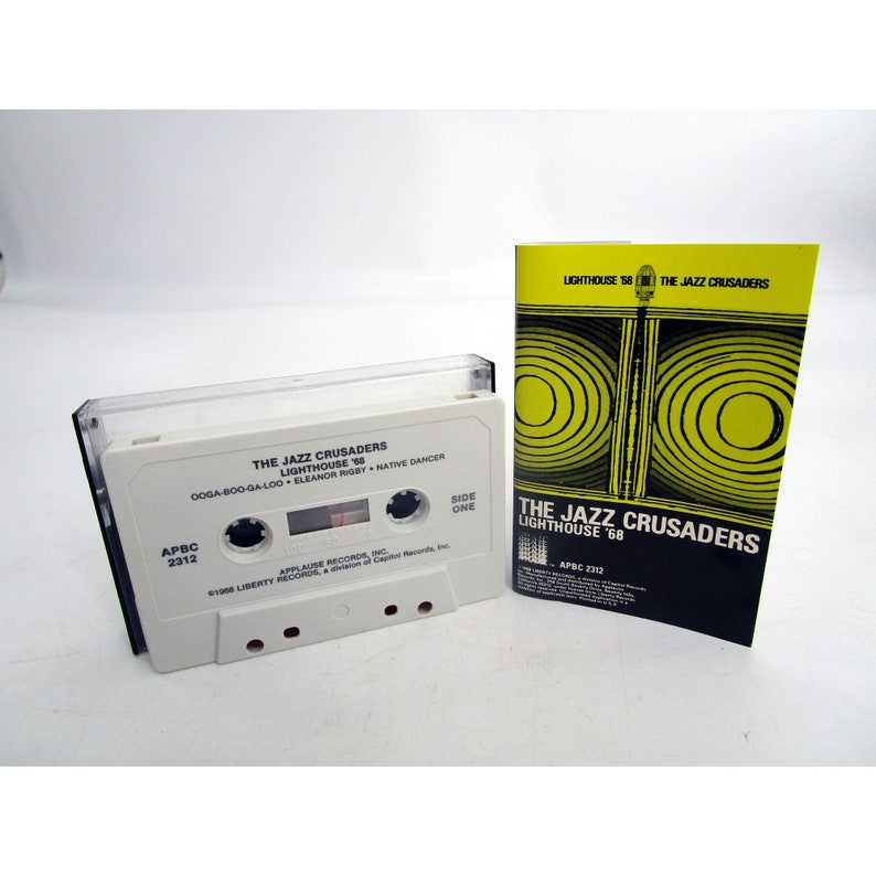 Op de afbeelding: Een witte cassetteband met de titel "The Jazz Crusaders Lighthouse '68" en de songtitels "Dooga-Boo-Ga-Loo", "Eleanor Rigby" en "Native Dancer". De cassetteband zit in een doorzichtige plastic hoes met een zwart label. De cassetteband ligt naast een zwart en geel papieren inlegvel met dezelfde titel en bandnaam.