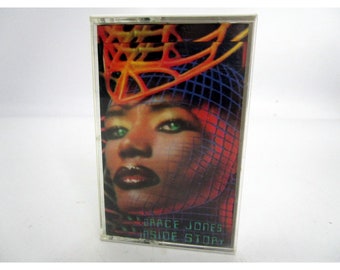 May include: A cassette tape with a colorful image of Grace Jones on the cover. The image features a woman with green eyes and red lips wearing a red, orange, and blue net headpiece. The text "GRACE JONES INSIDE STORY" is printed on the cover.