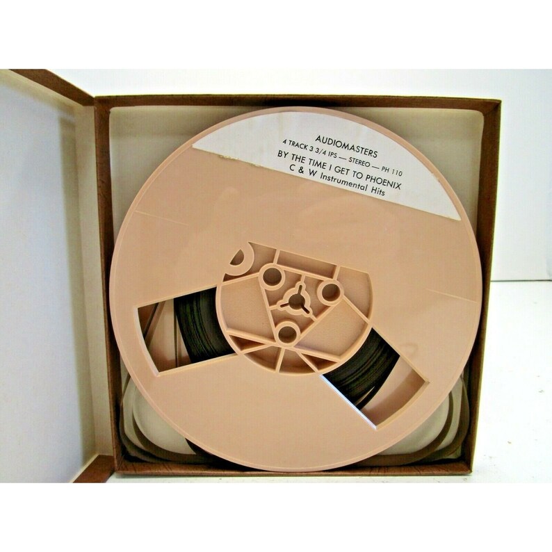 K&ouml;nnte beinhalten: Eine beige Audiobandrolle in einer braunen Box. Das Etikett auf der Rolle lautet "Audiomasters 4 Track 3 3/4 IPS Stereo - PH 110 By The Time I Get To Phoenix C & W Instrumental Hits".