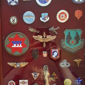 May include: A framed display of military badges and pins, including a US Air Force badge, a US Coast Guard badge, a US Marine Corps badge, and a US Navy badge. The badges are arranged in a grid pattern on a red background.