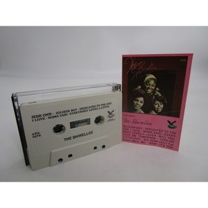 May include: A white cassette tape with a pink and white paper insert. The cassette tape has the text "The Shirelles" and "GTS-0079" printed on it. The paper insert has a photo of three women and the text "The Shirelles" and "GTS-0079". The text on the insert lists the songs on the cassette tape: "Soldier Boy", "Dedicated to the One I Love", "Mama Said", "Everybody Loves a Lover", "Will You Love Me Tomorrow", "Foolish Little Girl", "Big John", "(Ain't You Gonna Marry Mel)", and "Baby It's You".