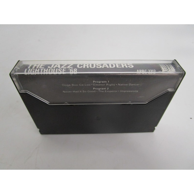 Op de afbeelding: Een zwarte cassetteband met de tekst "THE JAZZ CRUSADERS LIGHTHOUSE '68" en "ARRC 2317" gedrukt op de voorkant. De band vermeldt de nummers "Ooga-Boo Ga-Loo", "Eleanor Rigby", "Native Dancer", "Never Had It So Good", "The Emperor" en "Impressions".