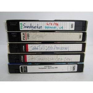 Pode incluir: Uma pilha de cinco fitas VHS com etiquetas escritas à mão. As etiquetas incluem os nomes de bandas e artistas como Soundgarden, Sting, Jethro Tull e Pat Travers. A fita de baixo tem o texto "Jack Bruce "Rockpalast" Alemanha 1980".