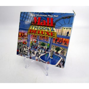 May include: A computer game box cover featuring a colorful illustration of a large shopping mall with a fountain in the center. The title of the game is "Mall Tycoon 2 Deluxe" and the text "Build The Ultimate Mega Mall" is at the top of the box.