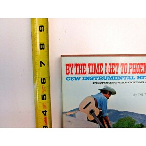 K&ouml;nnte beinhalten: Ein Vintage-Schallplattencover mit blauem Hintergrund und rotem und wei&szlig;em Text, der "By The Time I Get To Phoenix C&W Instrumental Hits Featuring The Guitar Of... By The..." lautet. Ein Mann mit Cowboyhut spielt auf dem Bild Gitarre.